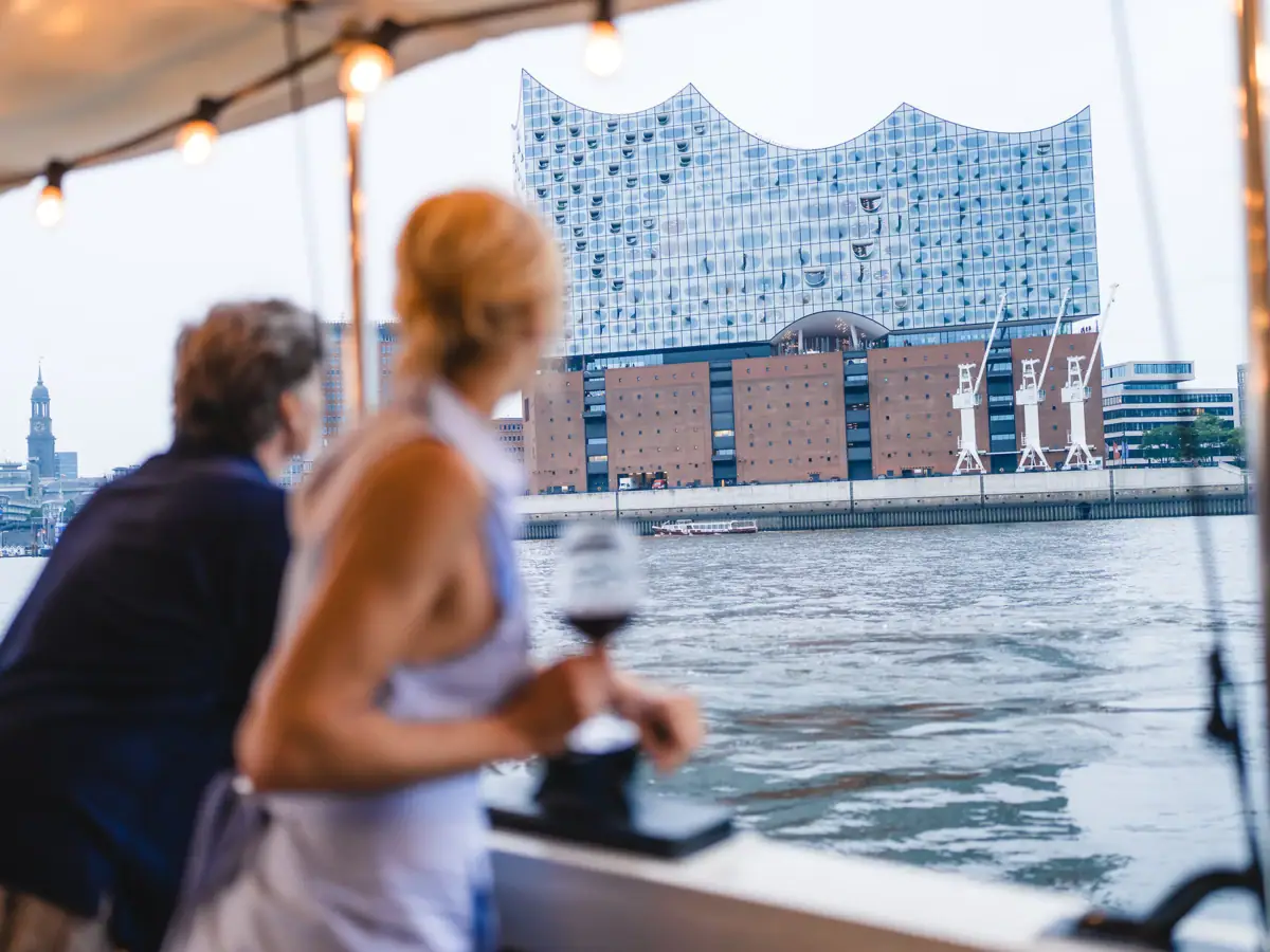 Ein Mann und eine Reling der Barkasse MS Jacob. Die Frau hält ein Glas Wein und schaut aus einem Boot auf die Elbe und die Elbphilharmonie.