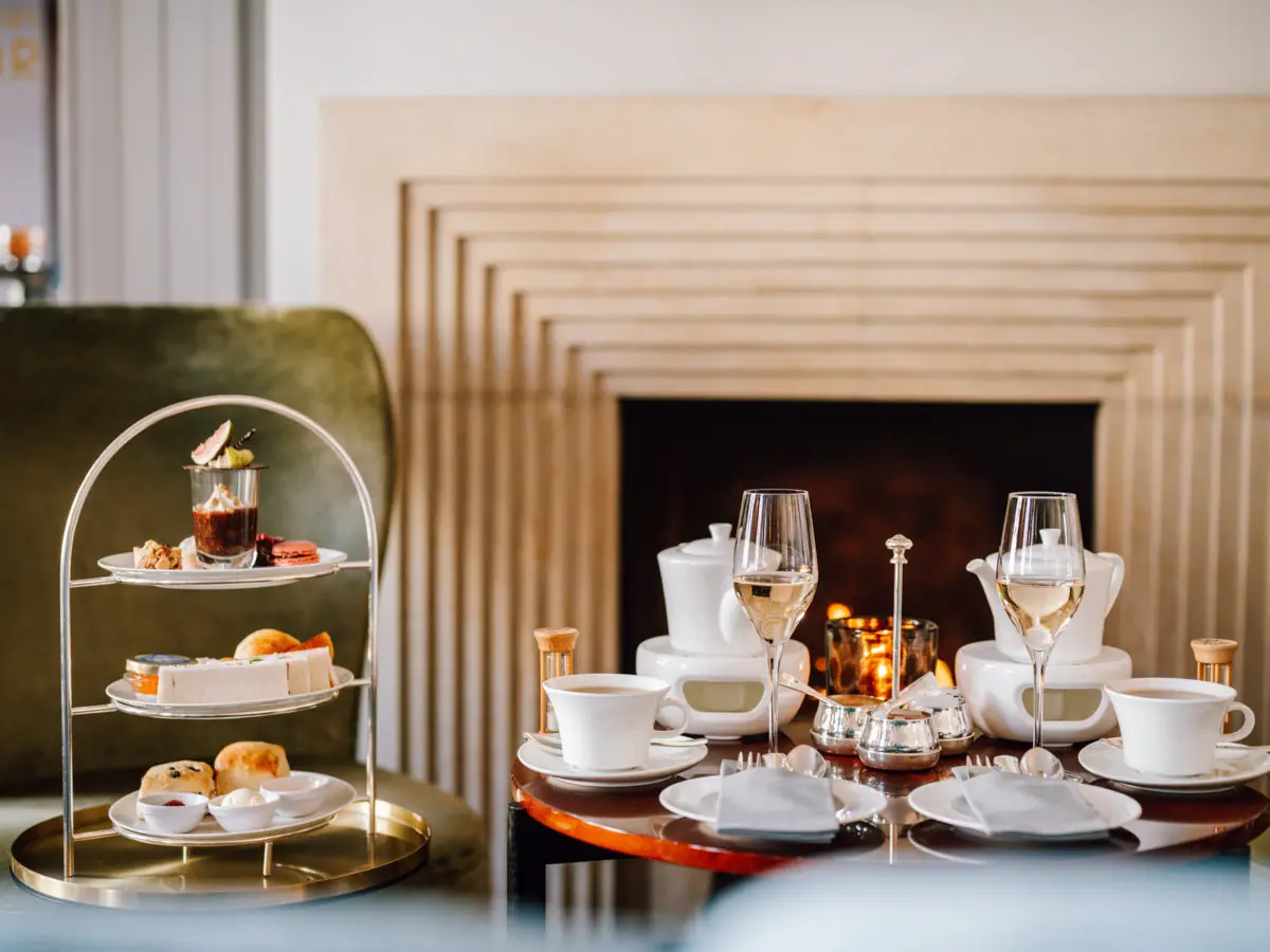 Teezeit im Hotel Louis C. Jacob. Vor dem Kamin in der Wohnhalle steht auf einem Tisch eine Etagere mit herzhaften und süßen Köstlichkeiten, Tee und zwei Gläsern mit Champagner.