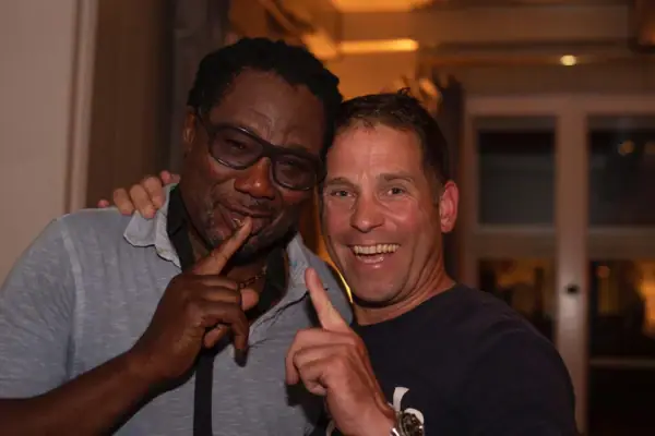 DJplus Combo Zwei Männer haben den Arm umeinander gelegt und halten den Zeigefinger hoch und lächeln in die Kamera.