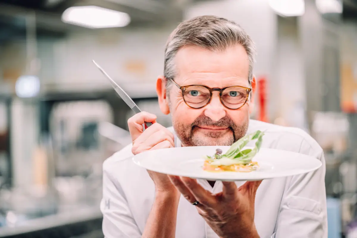 Chefkoch Thomas Martin hält in der einen Hand eine Pinzette und in der anderen hand einen Teller mit einem Gericht.