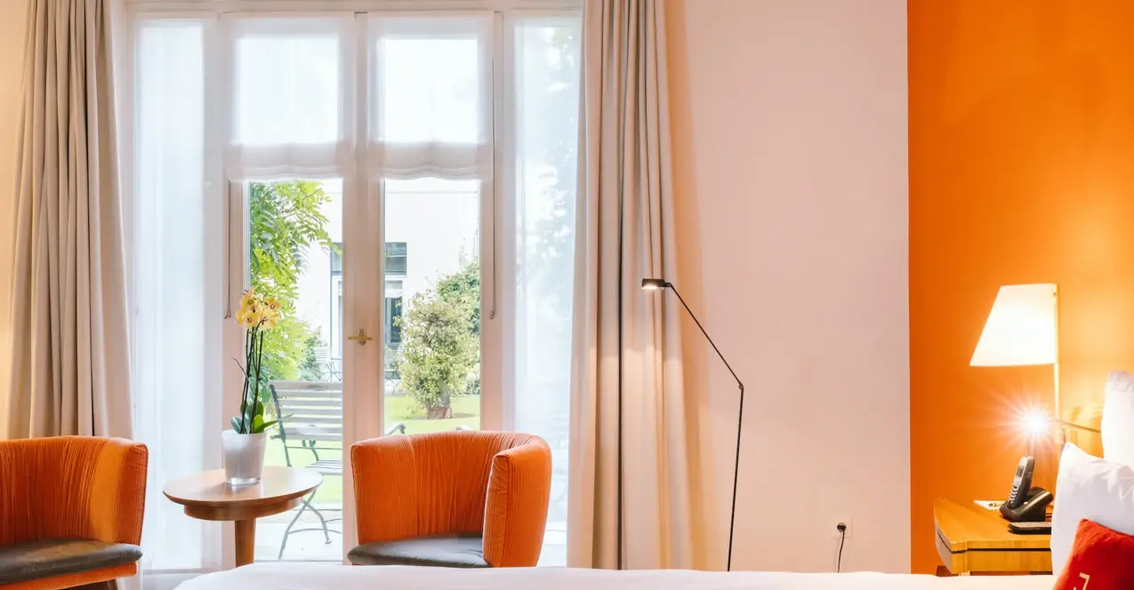 Vue intérieure d'une chambre d'hôtel au Louis C. Jacob avec un lit, une table, des chaises et une terrasse donnant sur le jardin.