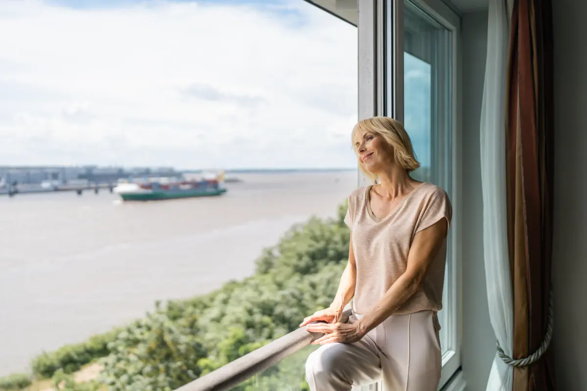 Eine blonde Frau schaut aus einem großen Fenster in einer Suite des Hotels Louis C. Jacob auf die Elbe und die fahrenden Schiffe.