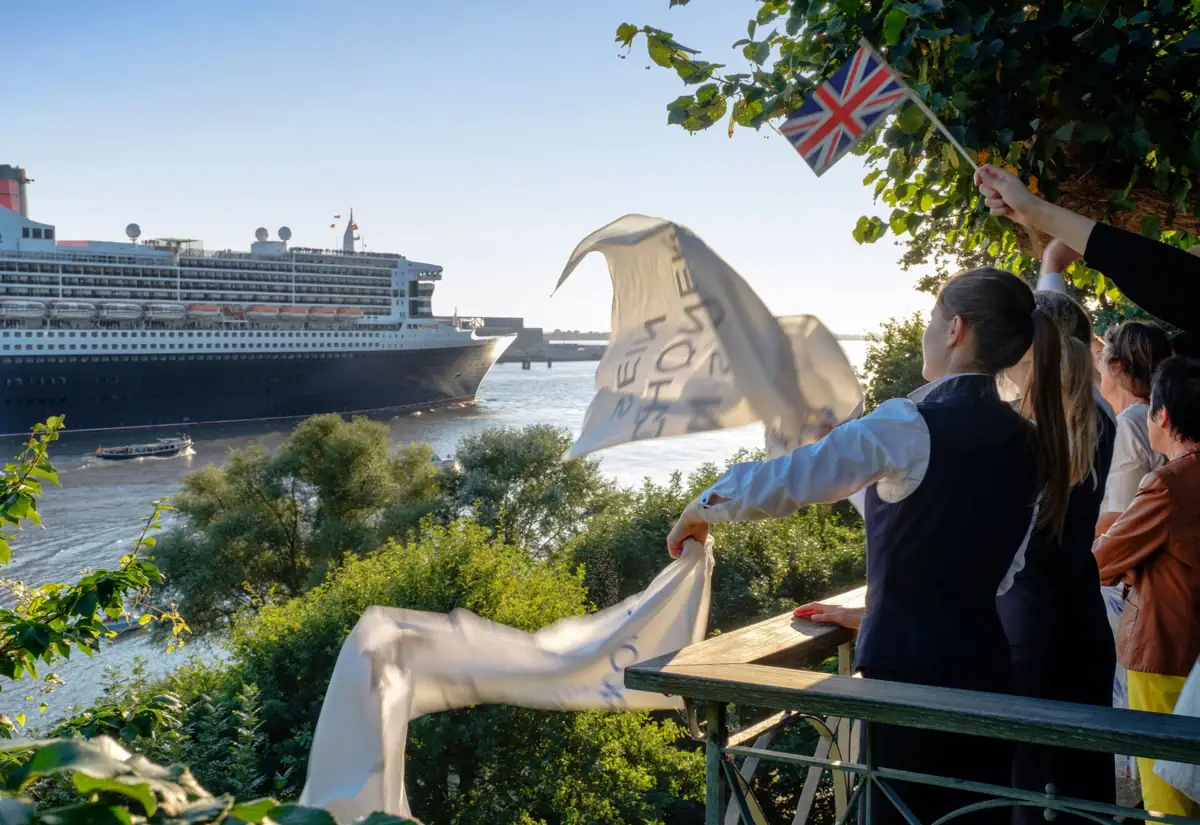 Lindenterrasse Mehrere Menschen schwenken Flaggen und verabschieden ein Schiff, welches auf der Elbe den Hamburger Hafen verlässt.