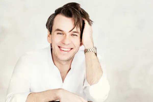 Eine Mann in weißem Hemd sitzt vor einem hellen Hintergrund und lacht in die Kamera. 