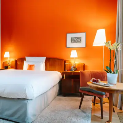 Zimmer mit orangener Wand, einem Bett, einem Stuhl und Beistelltisch im Hotel Louis C. Jacob.