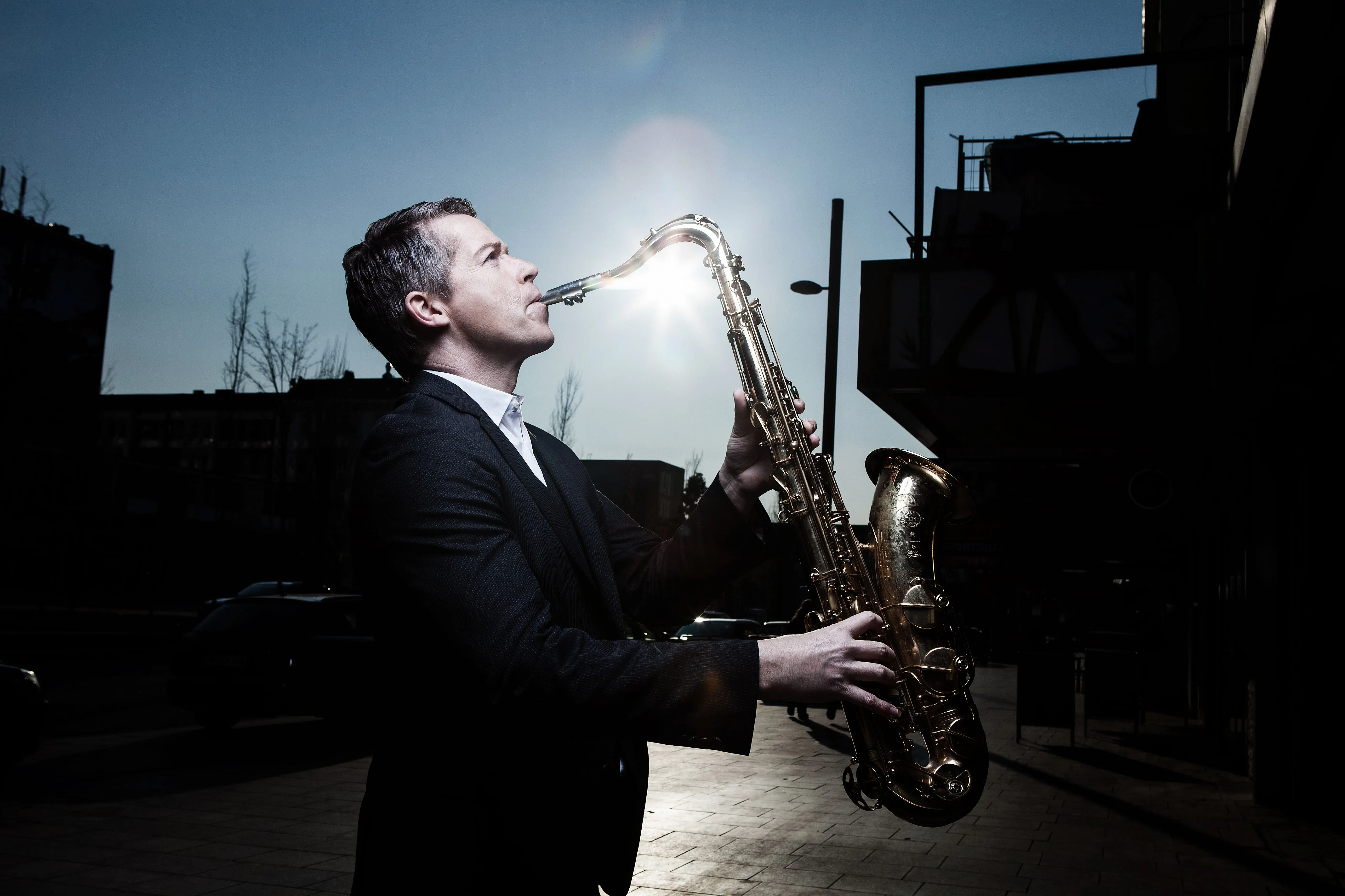 Toddi Reed Ein Mann in einem schwarzen Anzug spielt Saxophon und hinter ihm scheint die Sonne.