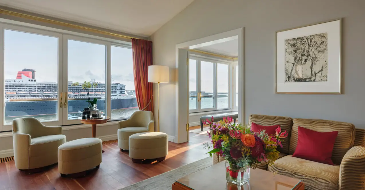 Salon de la suite Elbe de l'hôtel Louis C. Jacob avec canapé, fauteuils et table basse devant une grande fenêtre avec vue sur l'Elbe.