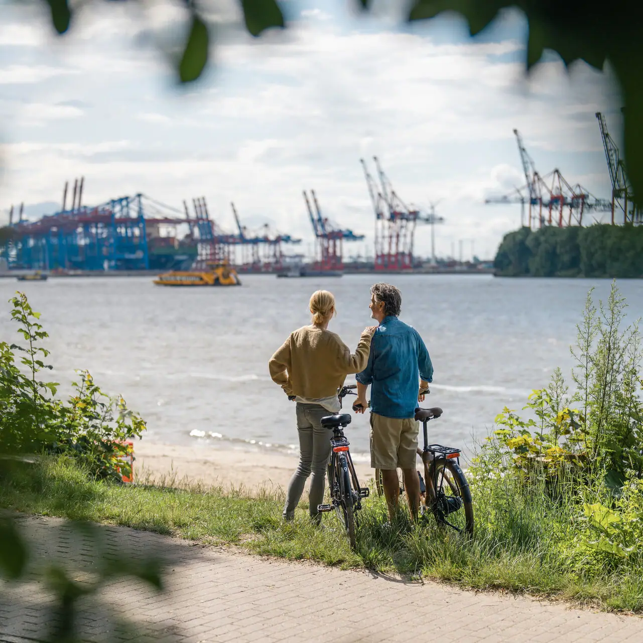 Fahrradtour Elbe Ein Mann und eine Frau stehen neben Fahrrädern an der Elbe und blicken auf den Hafen von Hamburg.