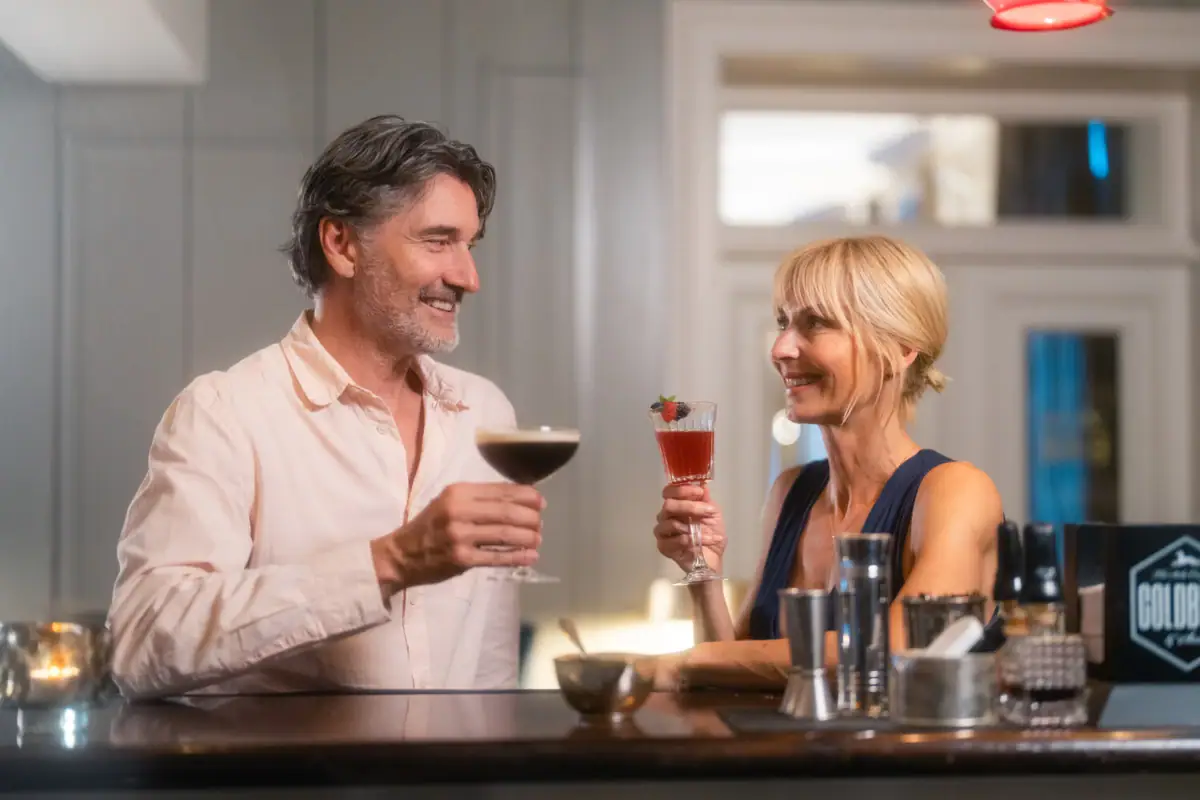 Eine blonde Frau und ein grauhaariger Mann stehen an der Jacobs Bar und halten je ein Cocktail-Glas in der Hand.