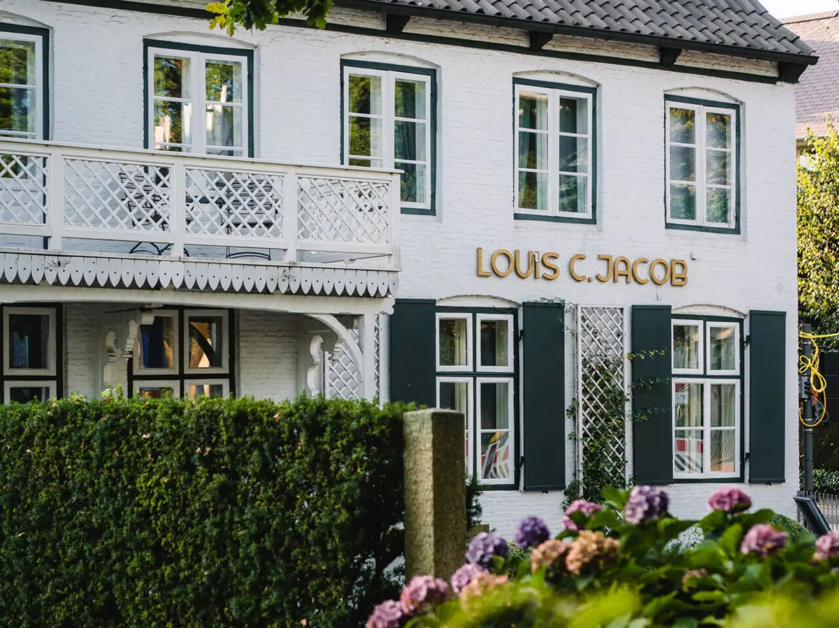 Weiße fassade des Hotels Louis C. Jacob mit einer grünen Hecke und lila Hortensienbüschen davor.