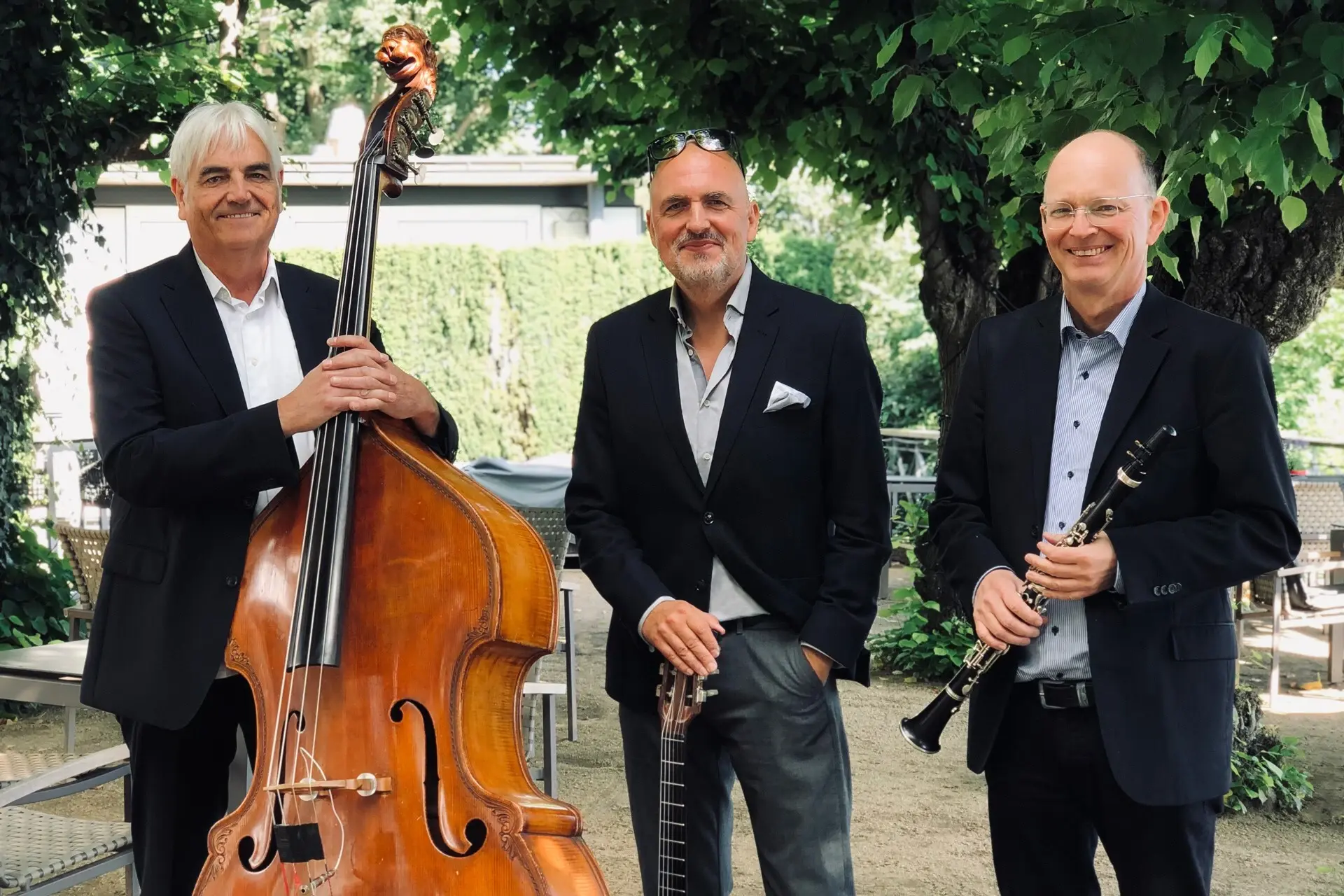 Hamburg String Brothers Drei ältere Männer halten jeweils ein Instrument, wie ein Cello, eine Gitarre und eine Oboe und lächeln in die Kamera.