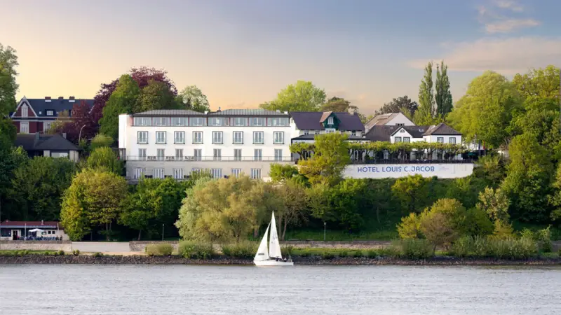 Das Louis C. Jacob Hotel Hamburg von außen mit einem Segelboot davor auf der Elbe.