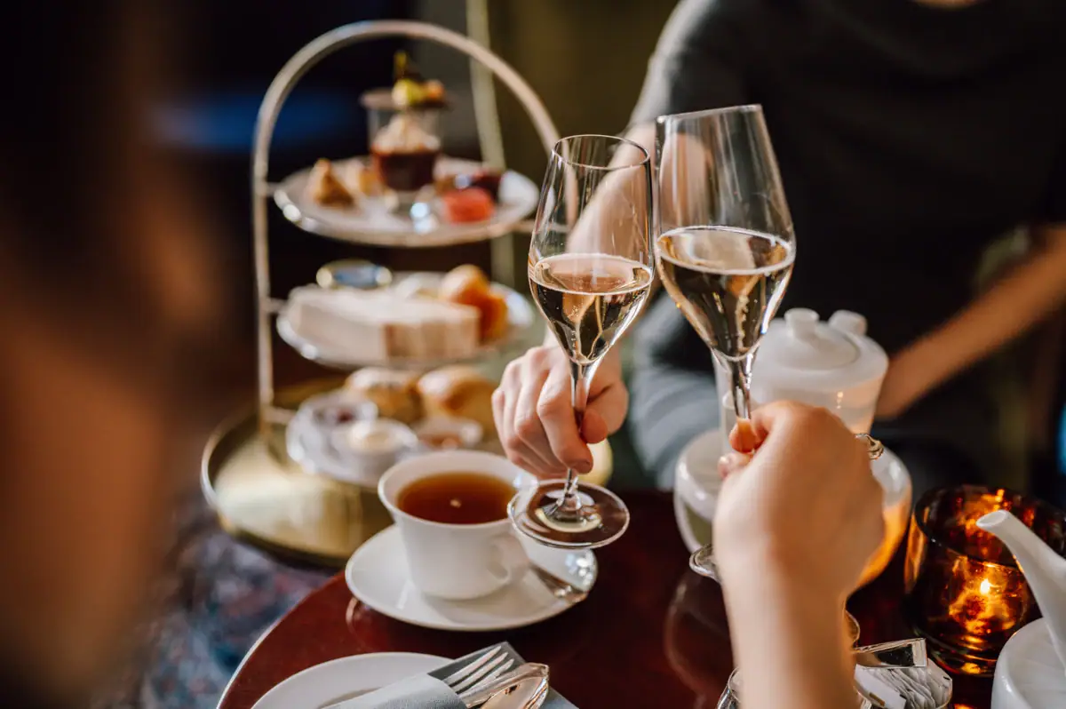 Teezeit im Hotel Louis C. Jacob. Zwei personen stoßen mit Champagner an und im Hintergrund sieht man eine Etagere mit herzhaften und süßen Köstlichkeiten.