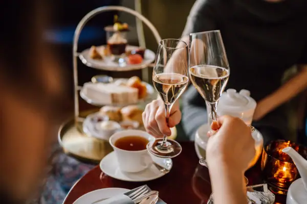 Afternoon Tea à l'hôtel Louis C. Jacob - une tasse de thé est posée sur la table et deux personnes trinquent au champagne. En arrière-plan, on aperçoit un plateau avec des scones, des sandwiches et des desserts sucrés.