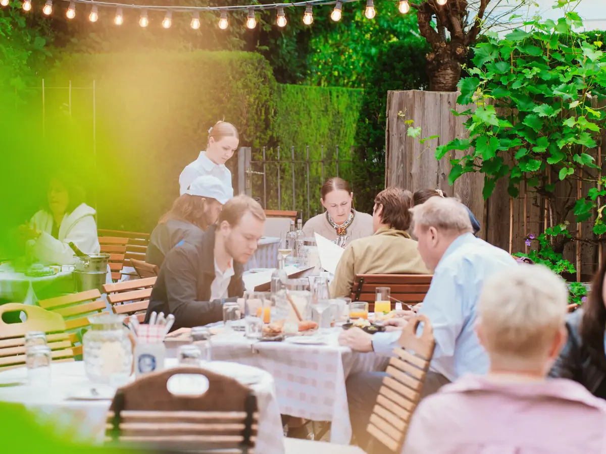 Personen sitzen an Holztischen im Biergarten der Weinwirtschaft Kleines Jacob. 